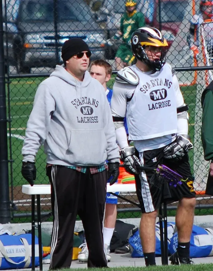 Two lacrosse players and a coach or spectator near a field fence.